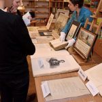 Osoba w białych rękawiczkach pokazuje dwóm osobom historyczne dokumenty i ilustracje w bibliotece. Stół jest wypełniony oprawionymi zdjęciami i otwartymi książkami. W tle widoczne są regały z książkami.